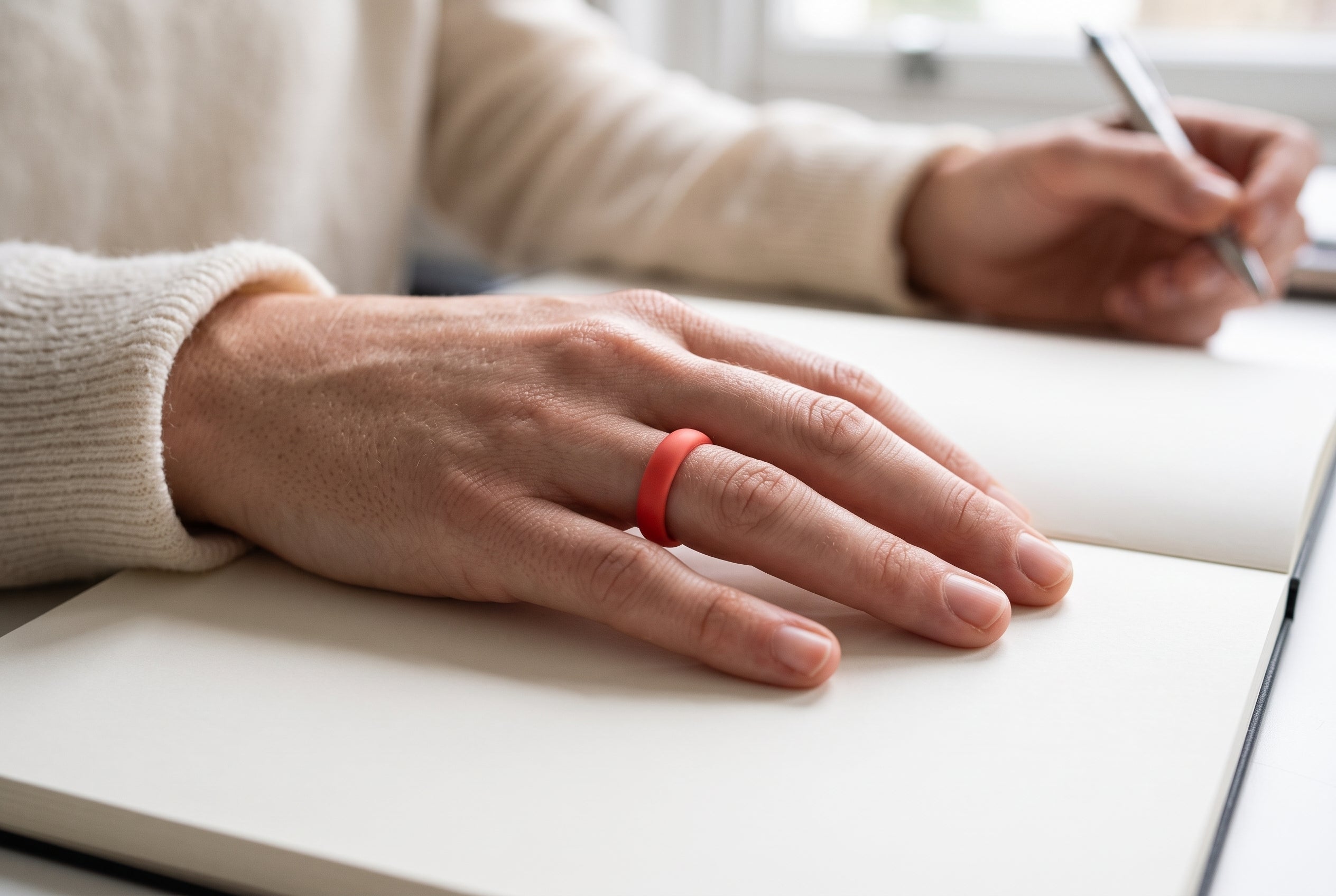 Red women's silicone ring worn with the hand resting on an open notebook at an office desk