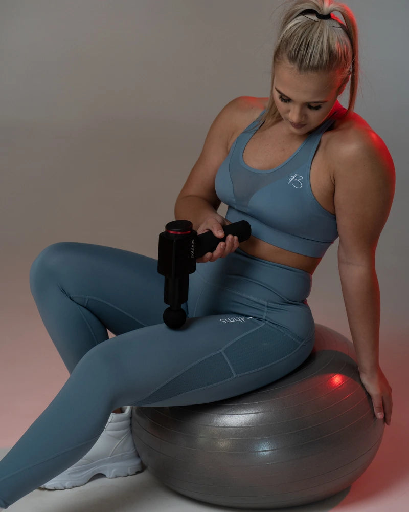 Young blonde trying botthms massage gun on her left leg muscle